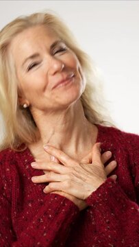 Vertical video of mature woman, receives beautiful compliments, finally raises her hands puts hands on chet. Beautiful mature blond isolated on solid white background. Copy space