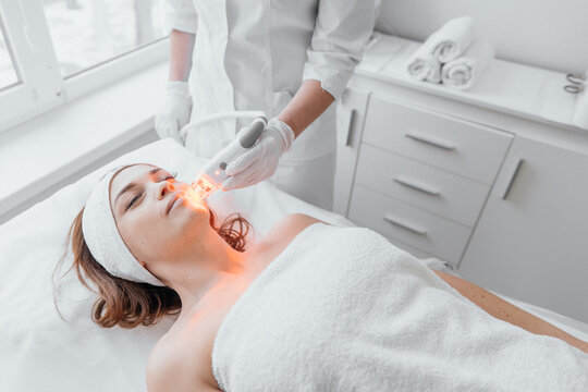 The beautician conducts light therapy of the face in a beauty clinic using an LED device. Procedure improves microcirculation and stimulates collagen production, and increases skin tone and elasticity
