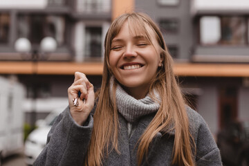 Young blonde with bang woman holds a key in her hand, standing on the street against new house...