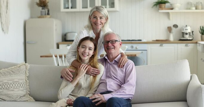 Happy couple of grandparent hugging teen grandkid, posing for shooting at home, looking at camera, smiling, laughing, sitting on couch, enjoying family leisure , granddaughter visit
