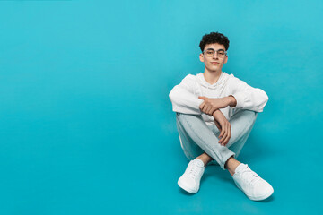 Confident teen boy in eyewear and white clothes, sitting down, looking at camera, isolated  turquoise background.