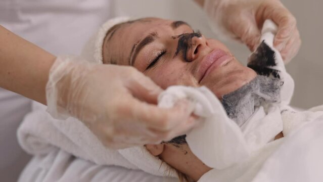 Beauty salon, Facial therapy, Mud therapy. With napkin, skincare specialist applies black mask on face of client at beauty salon, providing detox, lifting, kelp rejuvenation