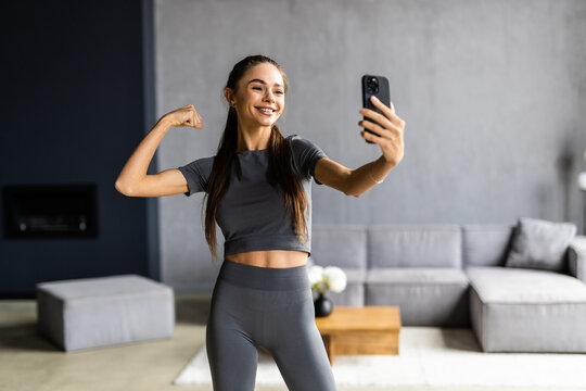 Portrait of sportive powerful woman dressed in tight gray top demonstrating her biceps taking selfie or having video call at home