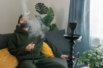 Bearded man is smoking hookah at home and blowing cloud of smoke - chill time and resting concept