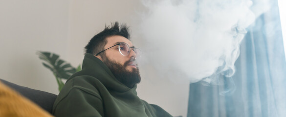 Banner bearded man is smoking hookah at home and blowing cloud of smoke copy space - chill time and...