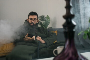 Bearded man is smoking hookah at home and blowing cloud of smoke - chill time and resting concept