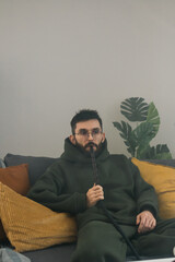 Obraz premium Bearded millennial or gen z man smoking hookah while relaxing on sofa at home - chill time and resting concept