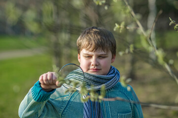A funny boy walks through the spring park, examines the leaves with a large magnifying glass, looks...