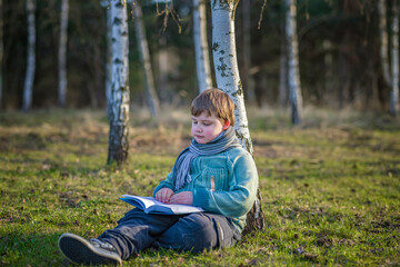A handsome boy in a scarf is in a spring park, sitting under a birch tree, reading a book and enjoying his dreams.