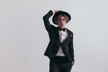 young elegant man in black tuxedo adjusting hat and posing with hand in pocket