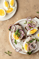 Smorrebrod sandwiches with herring fillet, rye bread, cream cheese, boiled egg slices, onion rings and microgreens on white dish brown background, rustic style. Top view