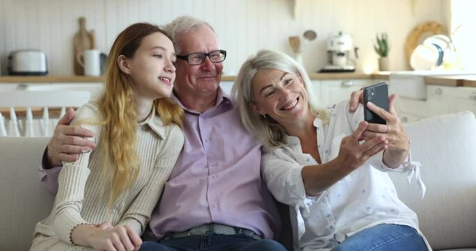 Cheerful couple of grandparents hugging teenage kid girl, resting on couch, taking selfie on cellphone, using media application for funny pictures, looking at screen, smiling, laughing, having fun