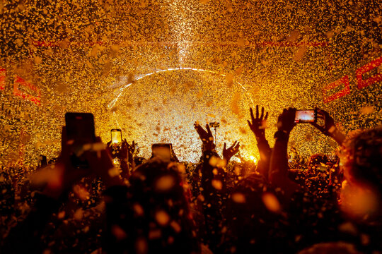 An energetic pop show in full swing! A crowd of fans is enthralled, their hands raised high, holding their phones to capture this party moment. Bright yellow lights and a flurry of confetti.