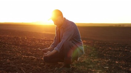 Agronomist touches soil examine and testing cultivated land before sowing on agricultural field and typing results in digital tablet on sunset. Smart farming technology and organic agriculture