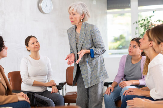 In study room, students sit in circle and sort out hypothetical task at work with teacher.Mature woman teacher stands next to students and invites them to participate in discussion