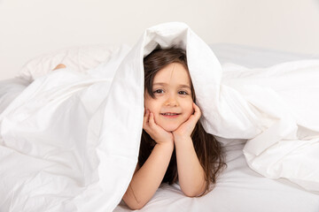 Fototapeta na wymiar Little attractive girl lying on bed and hiding under covers, she is smiling at camera. high quality