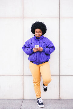 young latin afro woman standing on white wall using phone and smiling