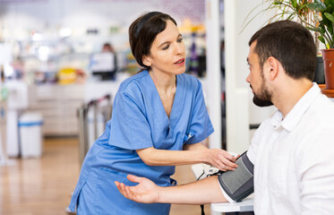 Obraz premium Portrait of positive female pharmacist checking blood pressure of male customer at pharmacy