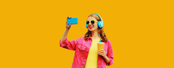Colorful portrait of happy cheerful laughing modern woman taking selfie with smartphone listening to music in wireless headphones wearing pink jacket on vivid yellow background - obrazy, fototapety, plakaty
