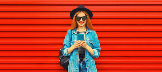 Portrait of stylish modern smiling young woman with smartphone wearing jean jacket, black round hat on red background