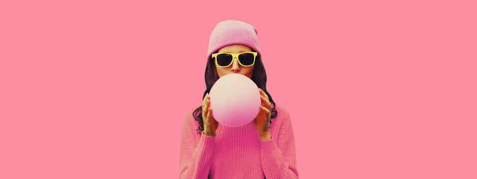 Fashionable colorful portrait of stylish cool young woman inflating chewing gum or balloon wearing hat on pink background