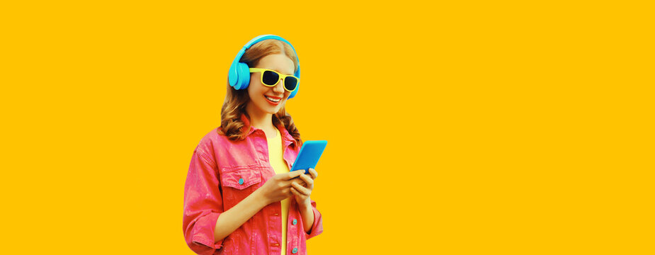 Portrait of stylish modern happy smiling young woman in headphones listening to music with smartphone wearing pink jacket on vivid yellow background