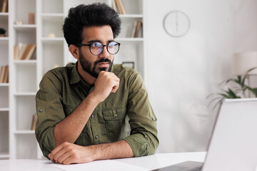 Bored millennial generation man sitting ay the table, feeling lonely at home. Unhappy young male suffering from negative thoughts, having depressive mood or problems in relations, regretting mistakes.