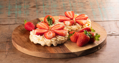 dessert of two waffles with white chocolate cream and pieces of strawberry on a round wooden board and cut strawberries on a wooden table close-up