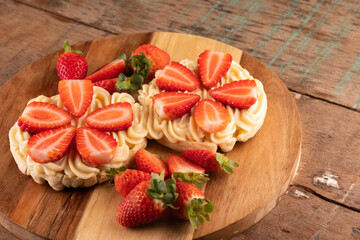 dessert of two waffles with white chocolate cream and pieces of strawberry on a round wooden board and cut strawberries on a wooden table close-up