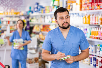 Friendly man pharmacist standing at drugstore with notepad and pen in hands