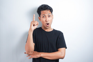 Young excited man in black t-shirt, pointing upwards with finger, having great idea, isolated on white background