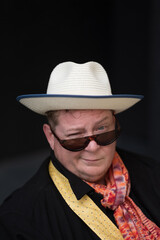 Wearing a scarf, loose tie, white hat, and fun sunglasses, a man in his powerchair expresses a wink and a smile, celebrating his presence as a disabled trans man.