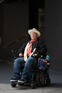 Outside of a stairwell, a man in a powerchair smiles confidently. Adding backstory, he is disabled and trans. His colorful scarf alludes to his LGBTQ identity.