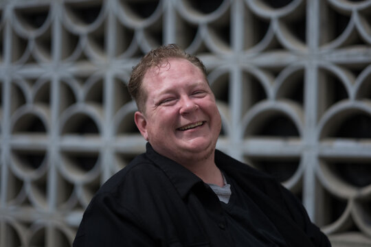 Posing in front of a cinder block wall, a man expresses confidence, joy, and pride in his identity as a disabled trans man living in the city.
