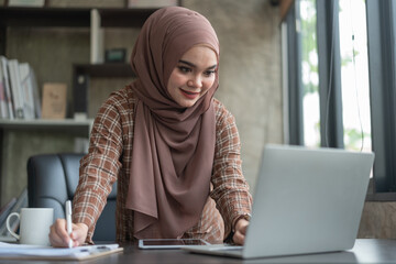 A beautiful Asian woman in hijab working from home looking at the screen to take notes in her book.
