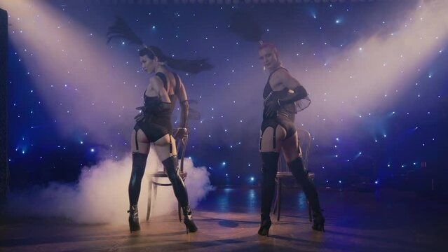 Two women in black underwear costumes sensually moving showing buttes, lowering down. Cabaret concept 4K. Sexy female burlesque dancers seducing client by erotic dance on theater stage or nightclub 