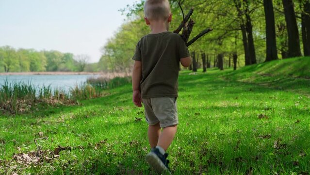 The camera follows a small boy walking across the lawn by the river. The child carries a bundle of firewood in his hands. The kid together with his family are relaxing in nature at the weekend.