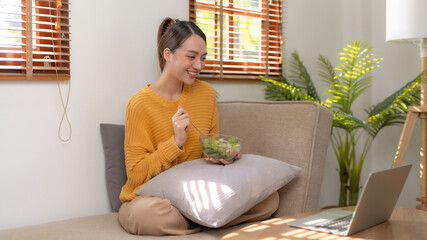 Happy young Asian woman eating salad at home and using laptop computer.