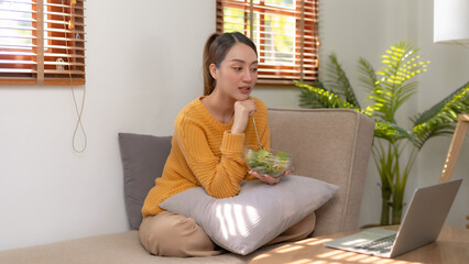 Happy young Asian woman eating salad at home and using laptop computer.