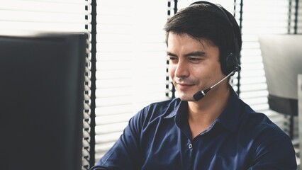 Young competent asian male call center agent working at his computer while simultaneously speaking...