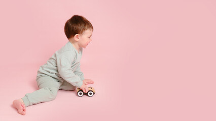 Happy toddler baby is playing with a toy car against a pink background. Child boy rolls a wooden toy car. Kid age one year eight months, full height