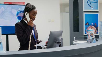 Young receptionist working at clinic reception counter, using landline phone to make checkup appointments with patients. Medical worker planning healthcare consultations at counter. Handheld shot. © DC Studio