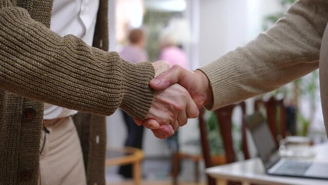 Close-up handshake of old Caucasian male friends indoors with blurred women at background talking. Unrecognizable men shaking hands meeting on weekend