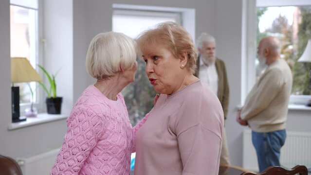 Senior Caucasian women gossiping about husbands standing indoors talking. Curious elderly lady listening friend whispering on ear sharing rumors. Femininity and eternal friendship