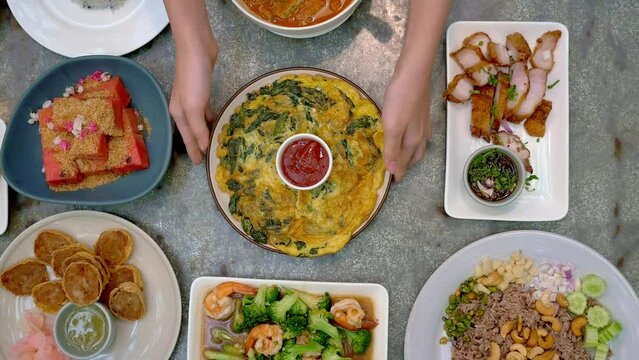 Top view of woman's hand serving Holy basil omelet among Thai food set, Grilled pork neck, Stir fried Broccoli, Watermelon with fish crispy, Shrimp paste rice. Asian cuisine food concept composition.