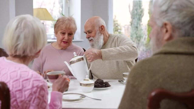 Caring senior husband pouring tea for loving wife sitting at dinner table thanking friends for visit. Portrait of confident Caucasian family talking with old friends dining in living room at home