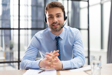 Cheerful young support phone male operator in headset, at workplace with collegues on the background while using computer, help service and client consulting call center concept.