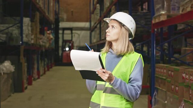 Concentrated female manager in white safety hardhat and vest taking notes on clipboard while checking availability of goods at large logistic storehouse. Manual inventory of supplies.