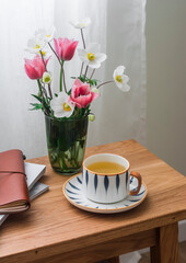A cup of green tea, a bouquet of spring garden flowers, a notebook on a wooden table in a cozy living room. Slow tea party