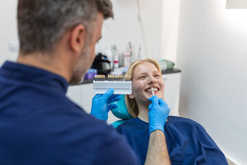 Image of satisfied young woman sitting in dental chair at medical center while professional doctor fixing her teeth, Female dentist choosing filling shade for smiling woman, using tooth scale sample